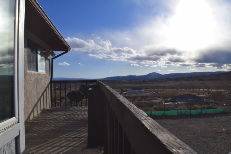 View from the upper deck to the west, Zuni Mountains in the skyline, blue sky, clouds coming in, bright sun. View overlooking the neighborhood to show fields and two homes.