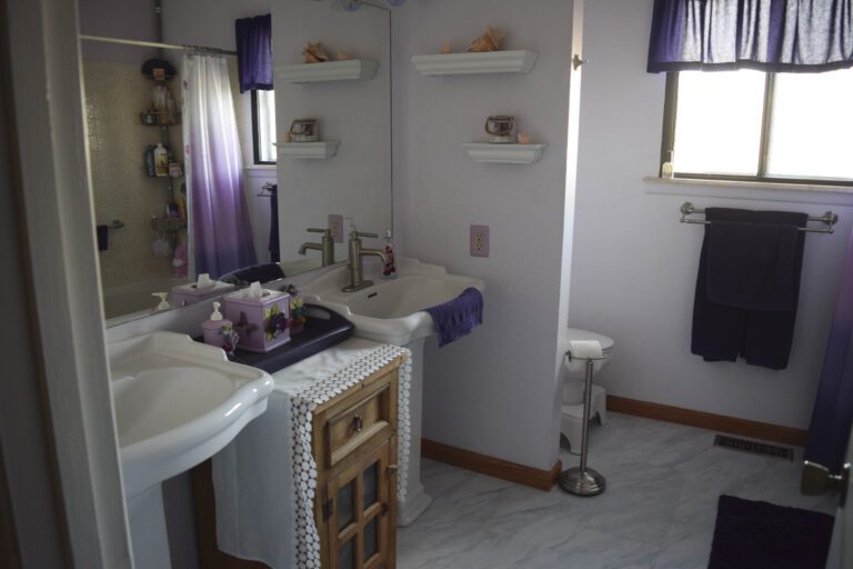 Bathroom to show white sink, drawer/vanity in the middle with mirror above, and another white sink to the right. To the far right partial wall and white commode. Right wall has a bath towel holder with a brown towel. Gray and white tile flooring