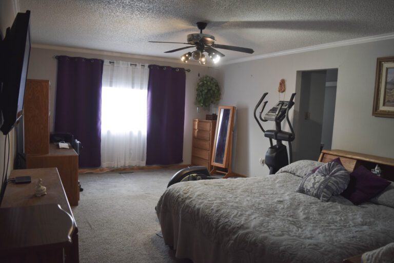 Bedroom with bed with gray comforter, walking treadmill along wall. Dresser and mirror stand in the corner. Large window with sheer window covering in the middle and purple curtains on each side open. TV mounted on left wall and dresser and desk along the same wall.