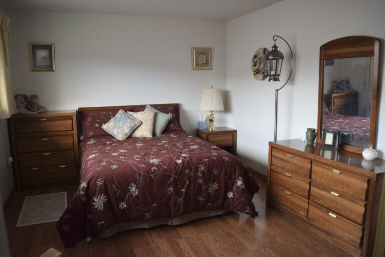 Bedroom with wood laminate flooring, nightstand with lamp with white lamp shade in the right corner of the room. Dresser with attached mirror along the wall to the right. Hanging lamp on a floor stand by the dresser. To the left of the bed is another brown wood 4 drawer dresser. Two pictures/paintings to the right and left of the wall in the background.