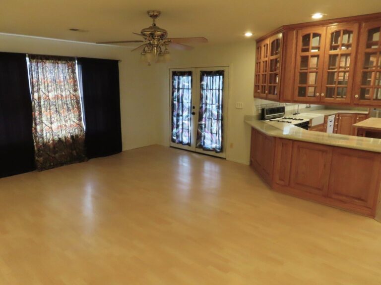 Spacious living second living area with ceiling fan and beige laminate wood flooring. Double doors with blue curtains in the background provide access to the backyard. Brown wooden Kitchen cabinets with glass pane doors, stove, and white counter tops in view to the right. Window to the left of the room with light blue and dark blue curtains.