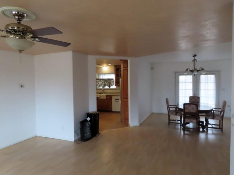 A spacious living area with polished beige laminate wood floors and a ceiling fan hanging from the ceiling. To the left is the entranceway to the kitchen. On the opposite end of the room is a dining table with 4 chairs and a double door exiting to the backyard.