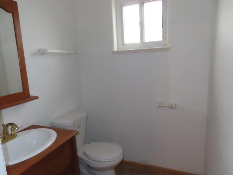 A compact bathroom featuring a white toilet, white sink, darker brown vanity, mirror over sink, and a smaller window.
