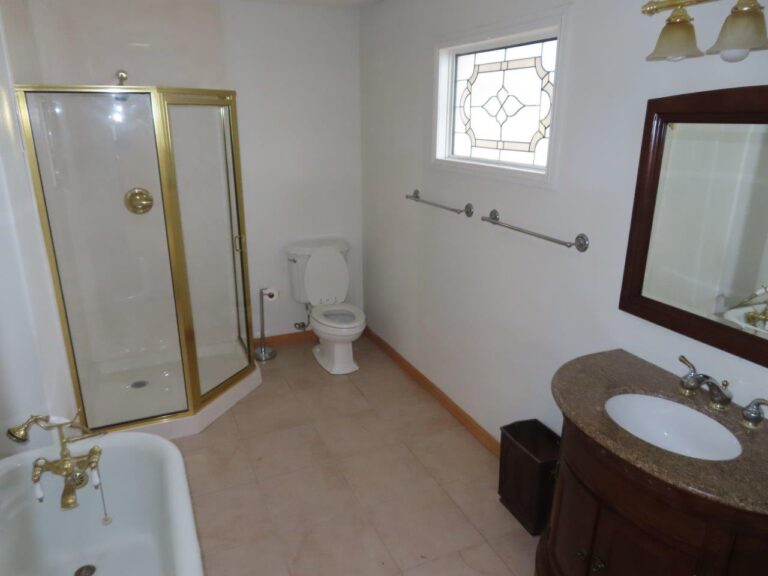 A large master bathroom with a clean, white toilet, a sleek dark brown colored sink with a mirror, stand-alone white tub, a spacious shower cubicle, and light brown tile. Art designed colored glass window on the right side wall.