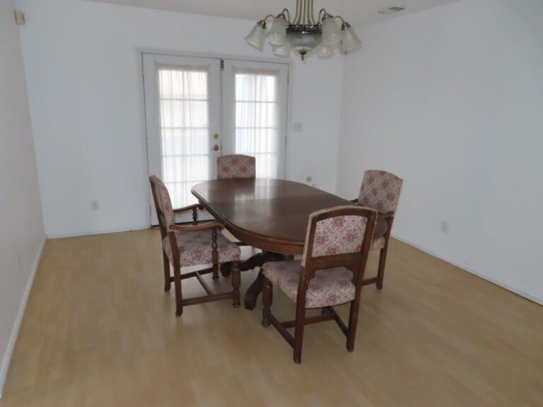 Dining area featuring a table and 4 chairs. Ceiling light over the table. Behind are wood and glass double doors exiting to the backyard. Light colored laminate wood flooring, white walls.
